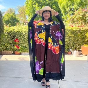 Pode incluir: Um kaftan longo e fluido, com um desenho de borboletas e flores em preto e castanho. O kaftan apresenta cores vibrantes, incluindo amarelo, laranja, vermelho e roxo. A pessoa est&aacute; a usar um chap&eacute;u de palha e sand&aacute;lias pretas.