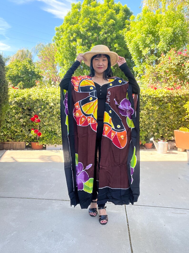 Pode incluir: Um kaftan longo e fluido, com um desenho de borboletas e flores em preto e castanho. O kaftan apresenta cores vibrantes, incluindo amarelo, laranja, vermelho e roxo. A pessoa est&aacute; a usar um chap&eacute;u de palha e sand&aacute;lias pretas.