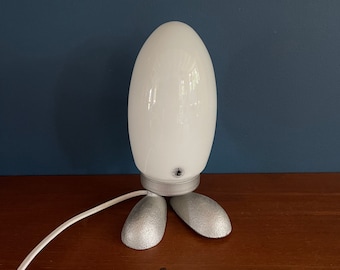 May include: A white ceramic table lamp with a rounded egg-shaped shade and three silver metal feet. The lamp has a white cord.