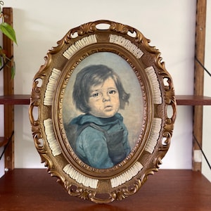 Puede incluir: Una pintura vintage ovalada enmarcada de un niño pequeño con cabello castaño y ojos azules, que lleva una camisa azul y un chaleco marrón, con una lágrima rodando por su mejilla. El marco es dorado con detalles blancos.