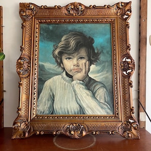 Puede incluir: Un retrato vintage de un niño en un marco dorado ornamentado. La pintura representa a un joven con una expresión pensativa, apoyando la barbilla en su mano. El fondo presenta un paisaje azul y blanco.