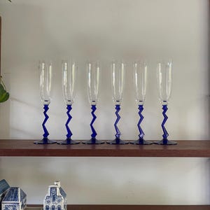 Puede incluir: Seis copas de champán de vidrio transparente con tallos en zigzag azul. Las copas están dispuestas en una fila en un estante de madera.