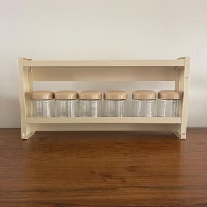 Op de afbeelding: Een beige kruidenrek met zes heldere glazen potten, elk met een bruine deksel. Het rek is gemaakt van een lichtbeige materiaal en is ontworpen om de potten in twee rijen te houden. Het rek staat op een houten oppervlak.