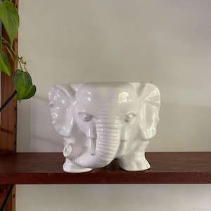May include: White ceramic elephant planter with a textured trunk and detailed features. The planter is sitting on a wooden shelf.