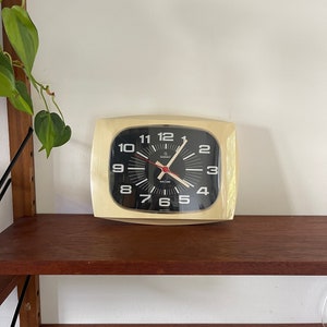 Puede incluir: Un reloj cuadrado de color crema con números y manecillas negros. El reloj tiene una esfera negra y una manecilla de segundos roja. El reloj está sentado en un estante de madera.