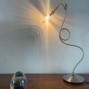 Könnte beinhalten: Eine moderne Schreibtischlampe aus gebürstetem Metall mit geschwungenem Design und rundem Fuß. Die Lampe ist eingeschaltet und wirft warmes Licht und Schatten an die Wand. Ein klarer Glasbriefbeschwerer mit dunklem Inneren liegt auf einer Holzoberfläche.