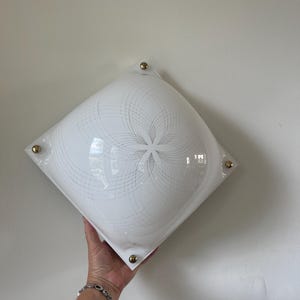 May include: A white, square ceiling light with a curved glass shade. The shade has a delicate, swirling grey pattern and is accented with four gold corner studs. The light is held by a person's hand. The fixture is a retro design.