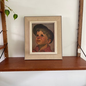 Puede incluir: Una pintura enmarcada de un niño pequeño con una gorra verde oscuro, mirando hacia arriba con una expresión esperanzadora. La pintura está sobre un fondo marrón claro y enmarcada en un marco de color crema.