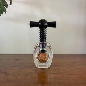 May include: Clear acrylic nutcracker with a black handle and a walnut inside. The nutcracker is on a wooden surface. The top of the nutcracker has a black handle. The walnut is visible through the clear acrylic.
