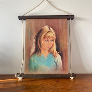 Puede incluir: Un retrato vintage de una persona joven con cabello rubio y ojos azules, vistiendo una blusa azul. La obra de arte se exhibe en un marco de madera con un colgador de cuerda. La firma del artista es visible en la esquina inferior izquierda.