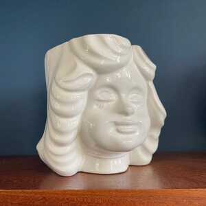 May include: White ceramic planter shaped like a woman's head with a stylized hairstyle. The planter has a closed mouth and closed eyes.