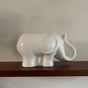 Può includere: Una statuetta di elefante in ceramica bianca con una proboscide e zanne. L'elefante è posizionato su una mensola di legno marrone scuro contro un muro grigio chiaro. La statuetta è un oggetto decorativo.