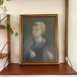 Puede incluir: Una impresión vintage enmarcada de un niño pequeño con un vestido azul oscuro, mirando hacia arriba con una expresión pensativa. La impresión está sobre un fondo oscuro y enmarcada en un marco dorado.