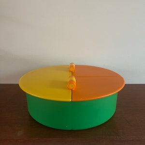 May include: A green, yellow, and orange round container with a lid. The lid has two orange and yellow knobs.
