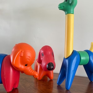 Puede incluir: Tres juguetes de animales de plástico de colores: un elefante naranja, un perro rojo y una jirafa con cabeza verde y cuello amarillo. El elefante tiene segmentos corporales azules, el perro tiene segmentos corporales naranjas y la jirafa tiene patas azules.