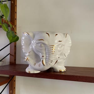 May include: White ceramic elephant planter with gold accents. The planter is sitting on a wooden shelf.