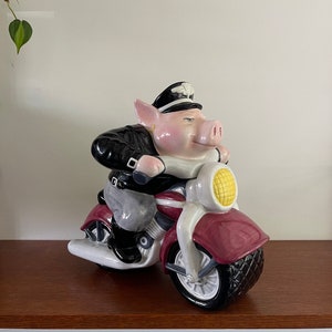Op de afbeelding: Een keramieken beeldje van een varken op een motorfiets. Het varken draagt een zwarte leren jas en een zwarte pet. De motorfiets is rood en wit met een gele koplamp. Het beeldje staat op een houten plank.
