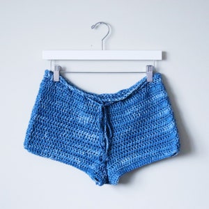 K&ouml;nnte beinhalten: Blaue H&auml;kelshorts, die an einem wei&szlig;en Kleiderb&uuml;gel h&auml;ngen. Die Shorts sind aus einem lockeren Strickstoff gefertigt und haben einen Bund mit Bindeband.