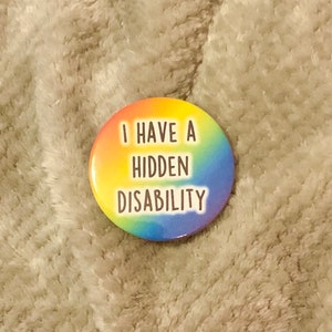Puede incluir: Un botón redondo con un fondo degradado de arcoíris. El texto blanco "I HAVE A HIDDEN DISABILITY" está centrado en letras en negrita. El botón está sobre un fondo suave y texturizado de color gris claro.