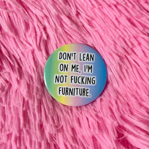 Może przedstawiać: Okrągła przypinka z tęczowym gradientem i napisem "DON'T LEAN ON ME, I'M NOT FUCKING FURNITURE." Przypinka jest umieszczona na różowym, puszystym tle.