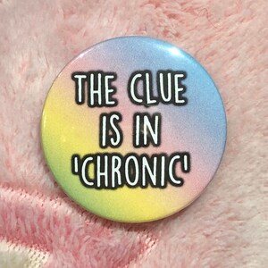Könnte beinhalten: Ein runder Button mit pastellfarbenem Regenbogen-Farbverlauf im Hintergrund. Der Text "THE CLUE IS IN 'CHRONIC'" ist in fetten schwarzen Buchstaben gedruckt. Der Button befindet sich vor einem weichen rosa Hintergrund.
