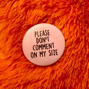 May include: A light pink button with the text "PLEASE DON'T COMMENT ON MY SIZE" in black, centered on the button. The button is set against a vibrant orange, fluffy background.