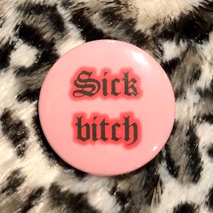 May include: A round, pink button with the words "Sick bitch" in black gothic font. The text has a red gradient effect. The button is set against a black and white animal print background.