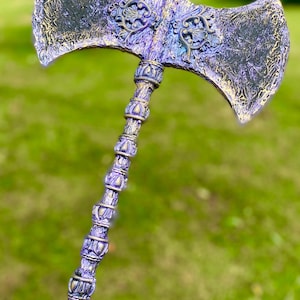 Puede incluir: Un hacha decorativa de doble hoja en morado y dorado. La cabeza del hacha presenta detalles ornamentados y una superficie texturizada. El mango también está intrincadamente diseñado. El fondo es un césped verde borroso. El hacha mide unos 76 cm de largo.