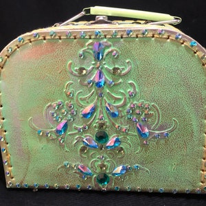 Puede incluir: Una caja decorativa verde de estilo vintage con asa. La caja está adornada con joyas iridiscentes y un ribete dorado. La parte delantera presenta un diseño floral en relieve con piedras preciosas azules y verdes. Un accesorio único y llamativo.