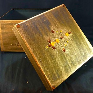 May include: A gold wooden box with a decorative floral design and amber accents. The box is open and the inside is lined with black velvet.