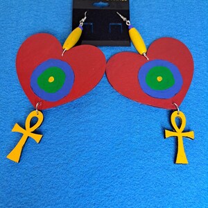 Puede incluir: Un par de pendientes de corazón rojo con un diseño circular azul, verde y amarillo. Cada pendiente tiene un amuleto de ankh dorado que cuelga de la parte inferior.