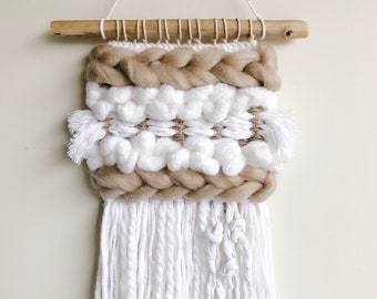 May include: A beige and white woven wall hanging with white fringe. The hanging has a braided design and a wooden dowel at the top.
