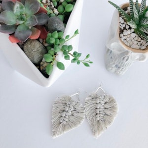 Puede incluir: Un par de pendientes de macramé hechos a mano de color gris claro con un diseño de flecos. Los pendientes cuelgan de ganchos plateados.