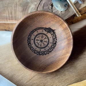 May include: A wooden bowl with a dark brown stain. The bowl has a circular design carved into the bottom, featuring a compass with runes and a snake wrapped around it.