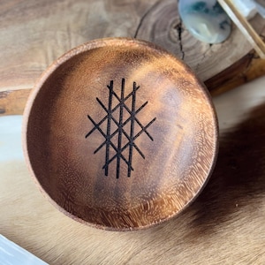 Op de afbeelding: Een houten kom met een donkerbruine vlek en een geometrisch ontwerp dat in de bodem is gegraveerd. Het ontwerp is een reeks kruisende lijnen die een raster vormen.