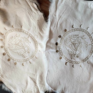Puede incluir: Dos pieles de cuero blancas con un dise&ntilde;o dorado de tres liebres en un c&iacute;rculo con un dise&ntilde;o de fases lunares alrededor.