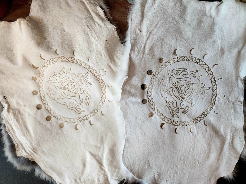 Puede incluir: Dos pieles de cuero blancas con un dise&ntilde;o dorado de tres liebres en un c&iacute;rculo con un dise&ntilde;o de fases lunares alrededor.
