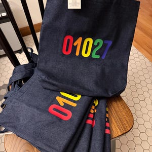 May include: Several dark blue denim tote bags with rainbow-colored "01027" text. The bags are on a wooden chair. One bag has a tag that says "Kinca Roo Art".