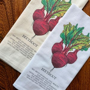 May include: Two kitchen towels, one cream and one white, each featuring a vibrant illustration of beetroots. The beetroots are depicted with deep red bulbs and green and yellow leaves. Text on each towel reads "BEETROOT" and a description of the beet's symbolism.
