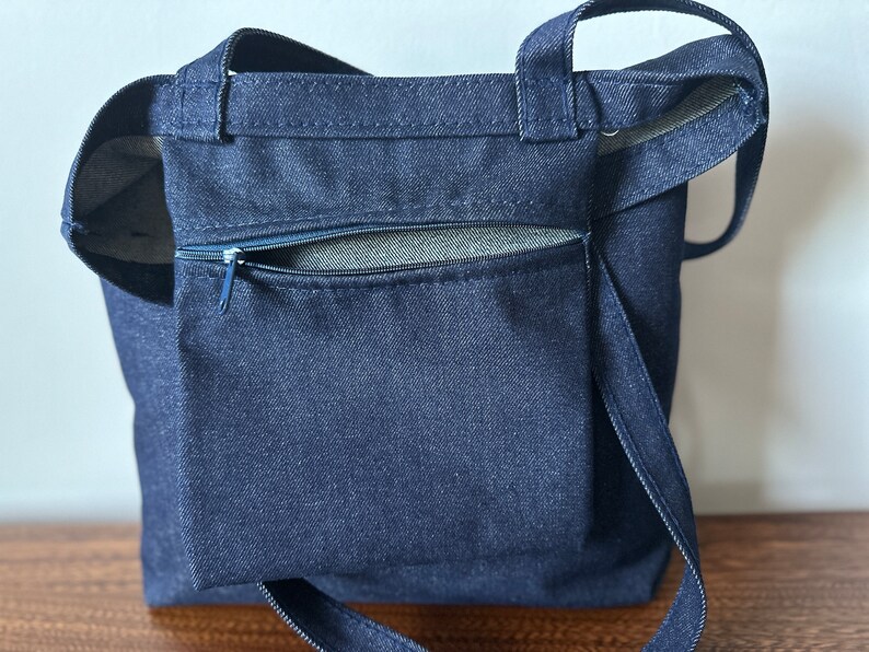 May include: A dark blue denim tote bag with a zippered pocket on the front. The bag has two long straps.