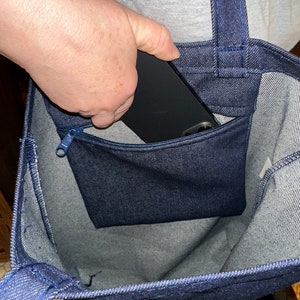 May include: A blue denim tote bag with a zippered pocket on the front. A black phone is being placed in the pocket.