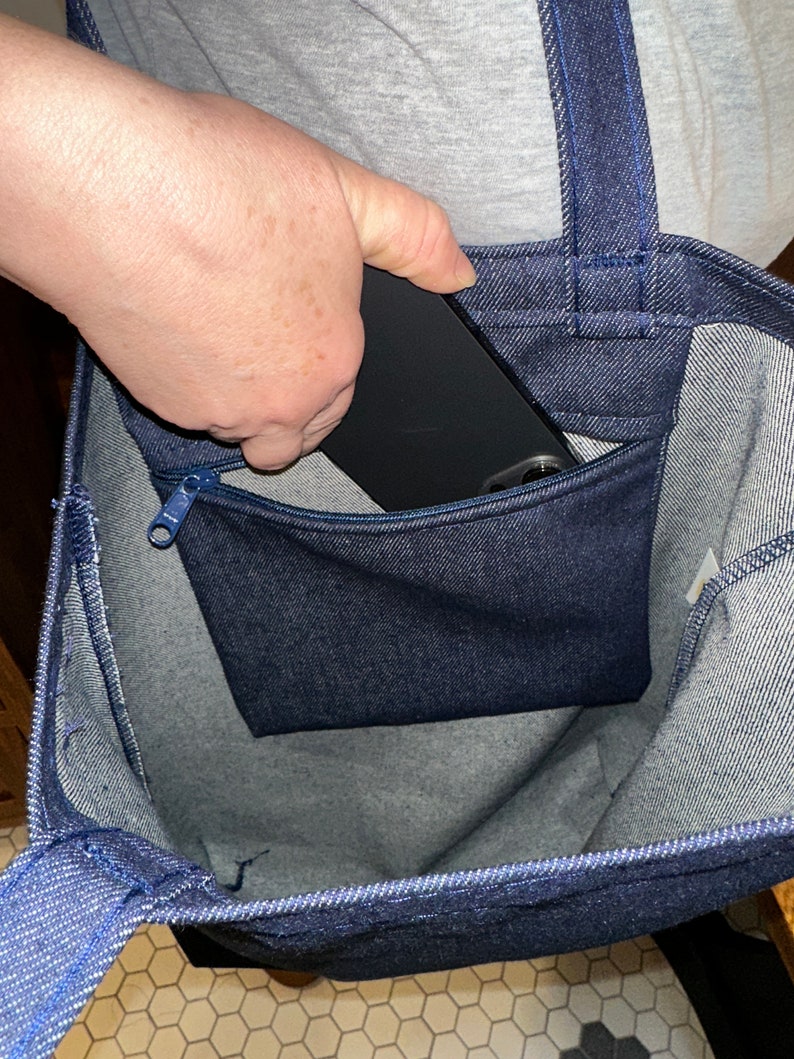 May include: A blue denim tote bag with a zippered pocket on the front. A black phone is being placed in the pocket.