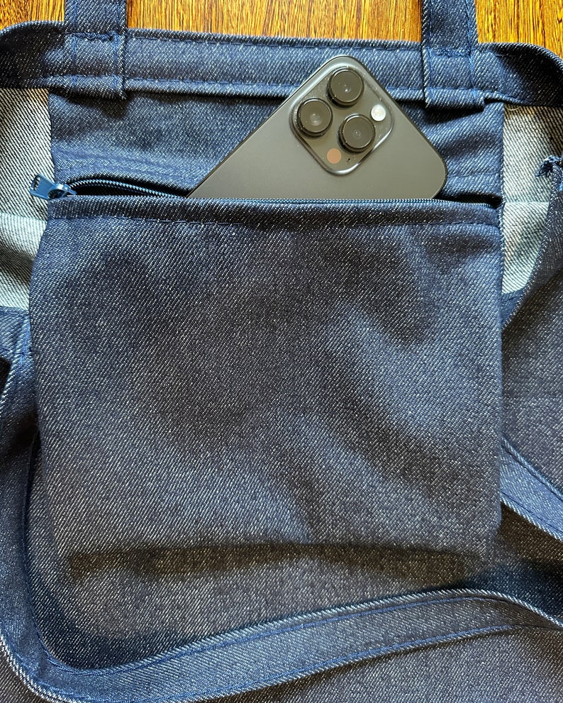 May include: A dark blue denim tote bag with a zippered pocket. A black smartphone with a camera lens is in the pocket.