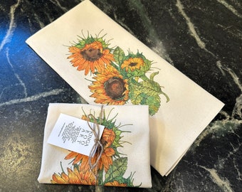 May include: Two tea towels with a sunflower design. The top towel has a large sunflower design with green leaves. The bottom towel has a smaller sunflower design with green leaves and the text "Kinga Aro" on a tag.