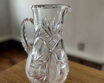 May include: A clear glass pitcher with a cut glass design. The pitcher has a handle and a spout.