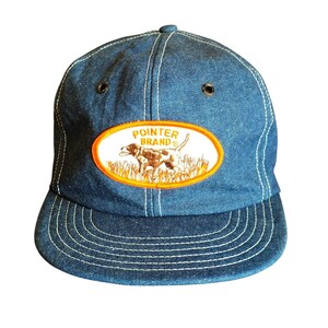 Op de afbeelding: Een blauwe baseballpet van denim met witte contraststiksels en metalen oogjes. Een ovaal embleem op de voorkant heeft een oranje rand, de tekst POINTER BRAND en een geborduurde afbeelding van een hond die in hoog gras staat.