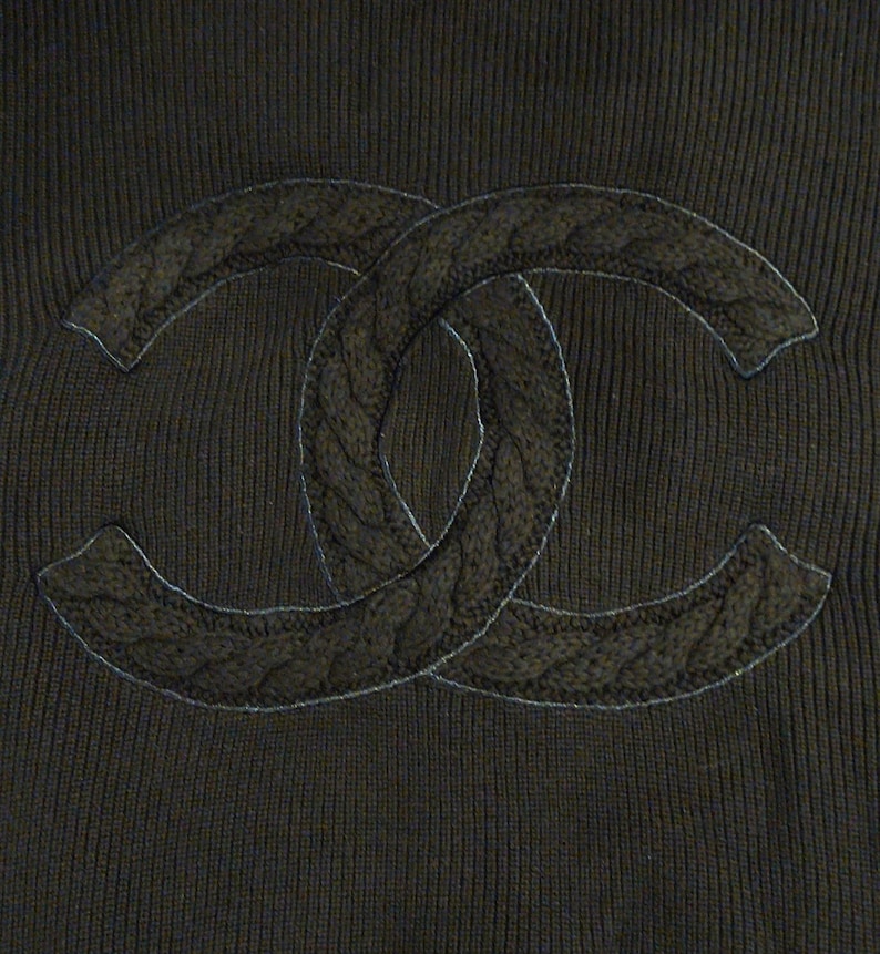 May include: Close-up of a dark brown knit fabric featuring a large, intertwined double-C logo. The logo is textured with a cable knit pattern and outlined with a thin, light-colored thread, creating a subtle contrast against the darker background.
