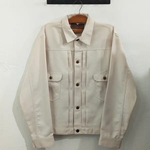 May include: A cream-coloured button-up jacket featuring a classic collar, long sleeves, and two chest pockets with buttoned flaps. The front of the jacket displays vertical pleat detailing along the placket, secured with brown buttons.