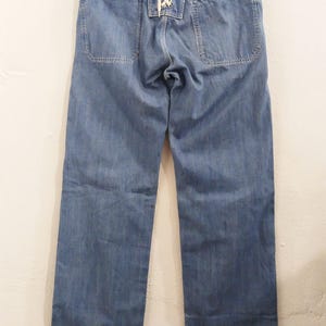 May include: Pale blue denim jeans with a distinctive lace-up feature at the rear. The jeans have a traditional five-pocket design and a straight leg cut. The jeans are suspended from a black hanger.
