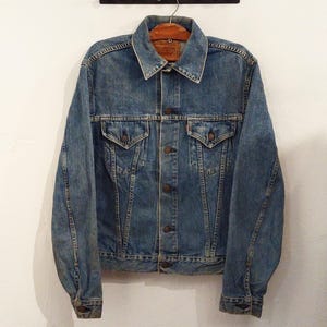 May include: A classic denim jacket in a medium blue wash. The jacket has a collar, button closure, two chest pockets, and buttoned cuffs. It is suspended on a wooden hanger against a white wall.